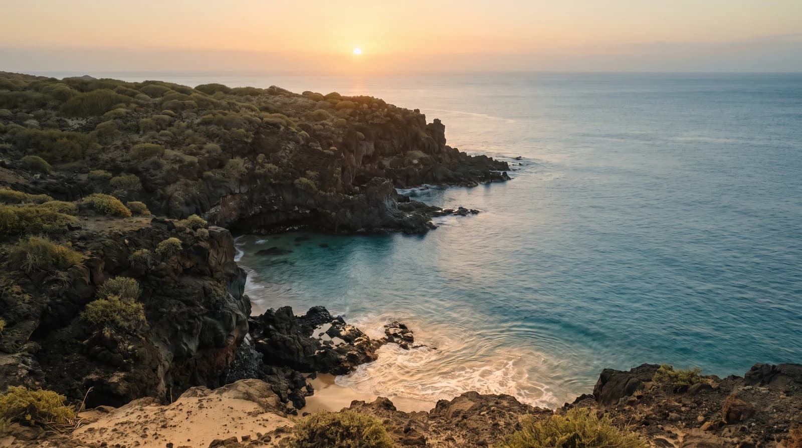 Rugged volcanic coastline at golden hour, with a sheltered turquoise cove below olive-green headland scrub