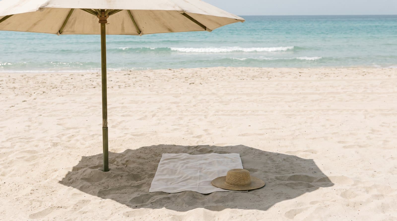 A folded towel and a straw hat in the shade of a canvas beach umbrella, with the sea beyond.