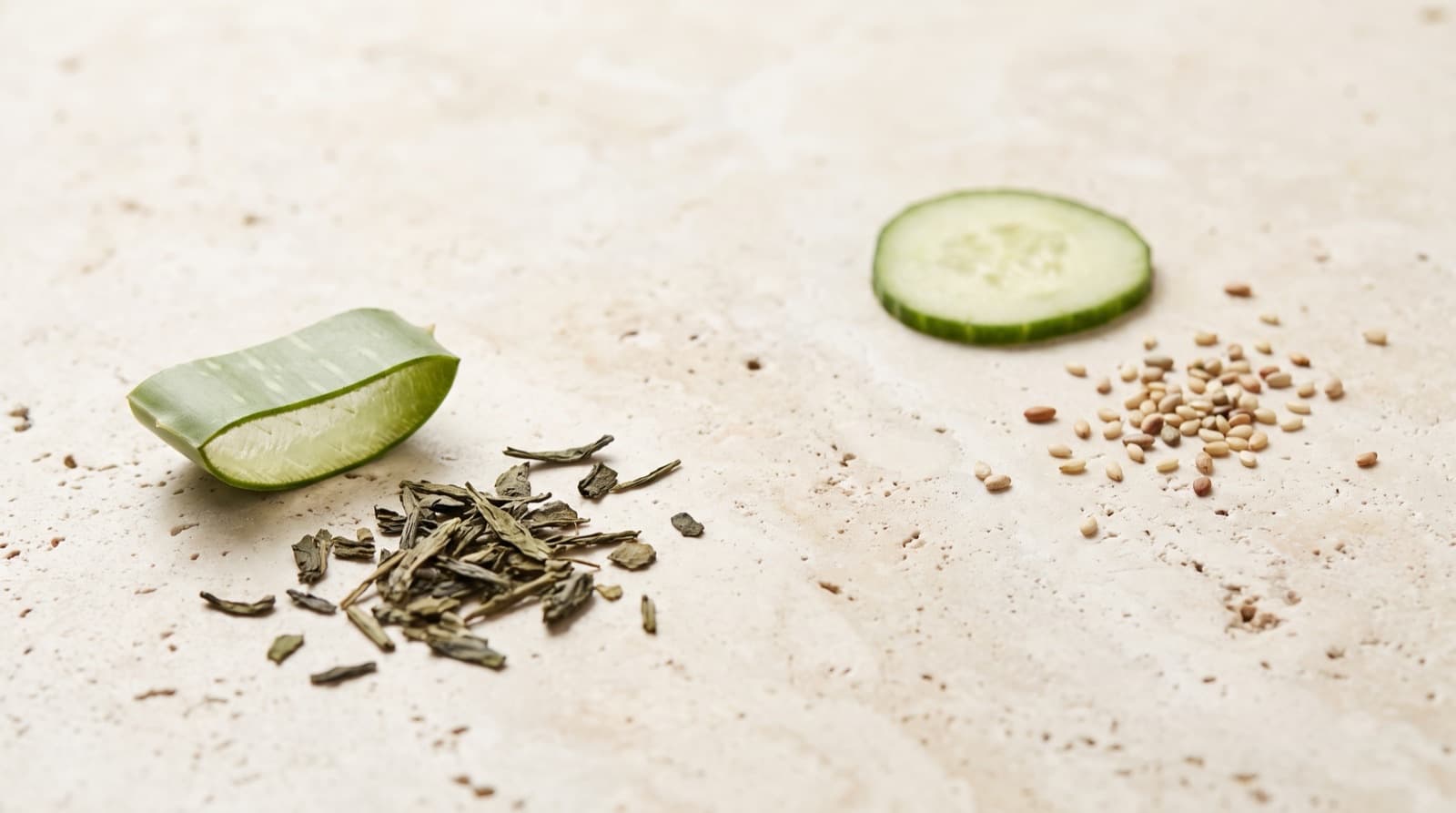 A cut aloe leaf, loose green tea leaves, a cucumber slice and a scatter of pale seeds, laid separately on pale limestone.