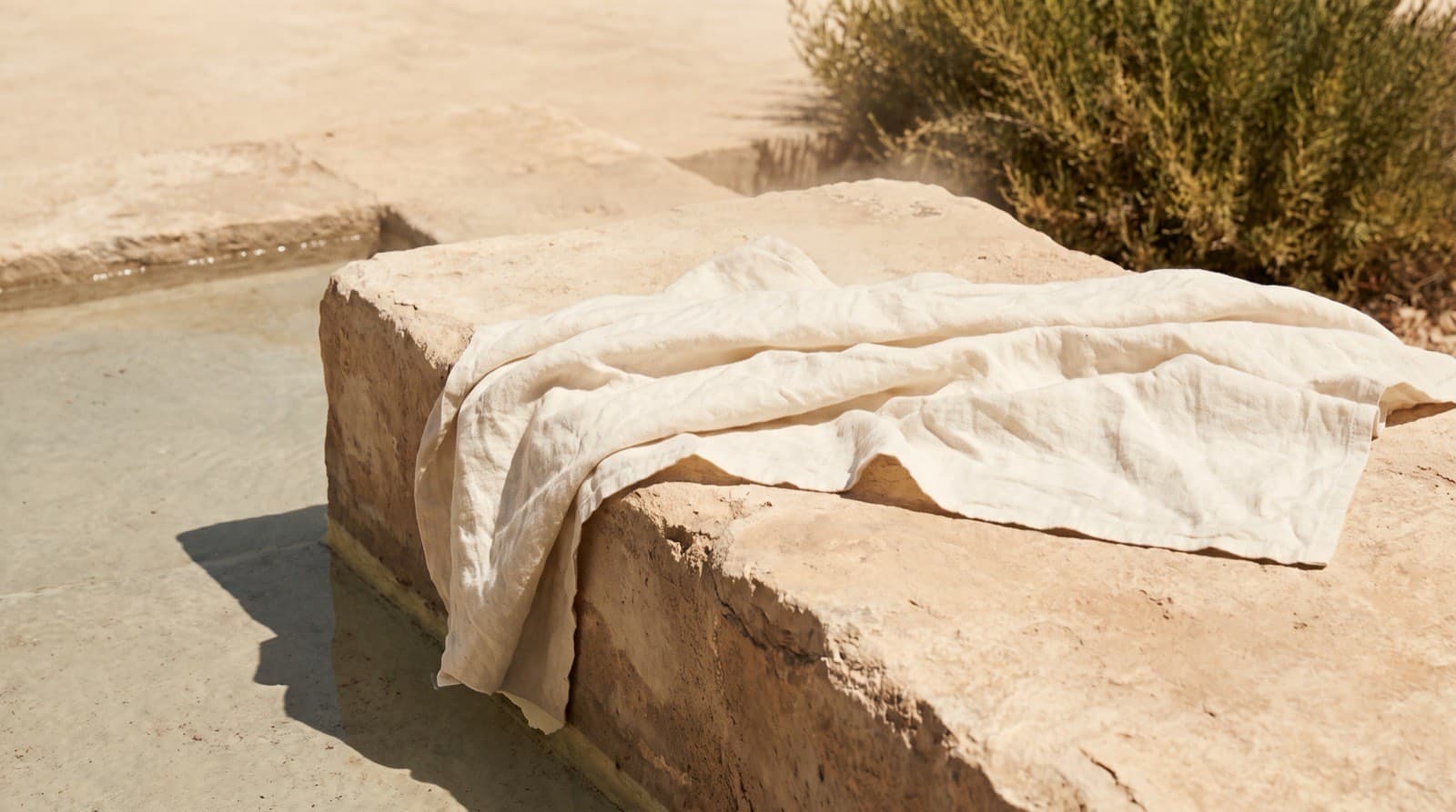 Ivory linen draped over warm stone in hard overhead midday sun, beside a shallow ledge of still water.