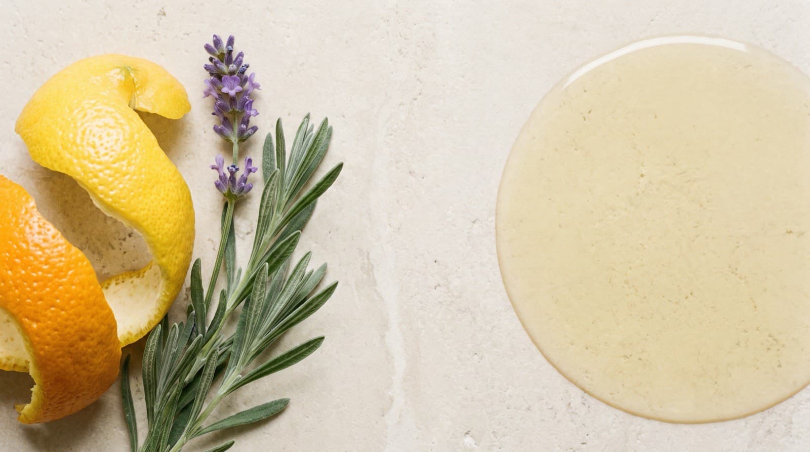 Citrus peel and a lavender sprig on one side of a pale stone surface, and a still pool of pale golden oil on the other.