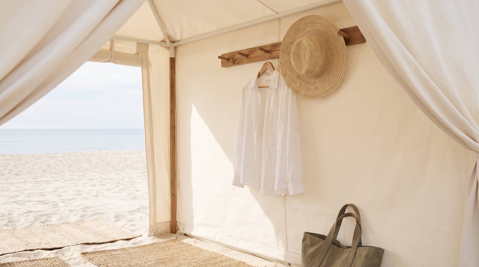 A long-sleeved white linen shirt and a wide-brimmed straw hat hanging from a wooden peg in the shade of a canvas beach cabana, with sand and sea beyond.