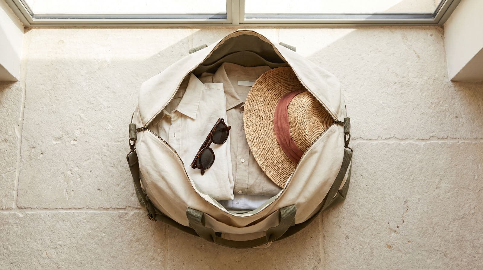 An open canvas weekender bag on a stone floor, packed with folded linen shirts, a straw hat and sunglasses.
