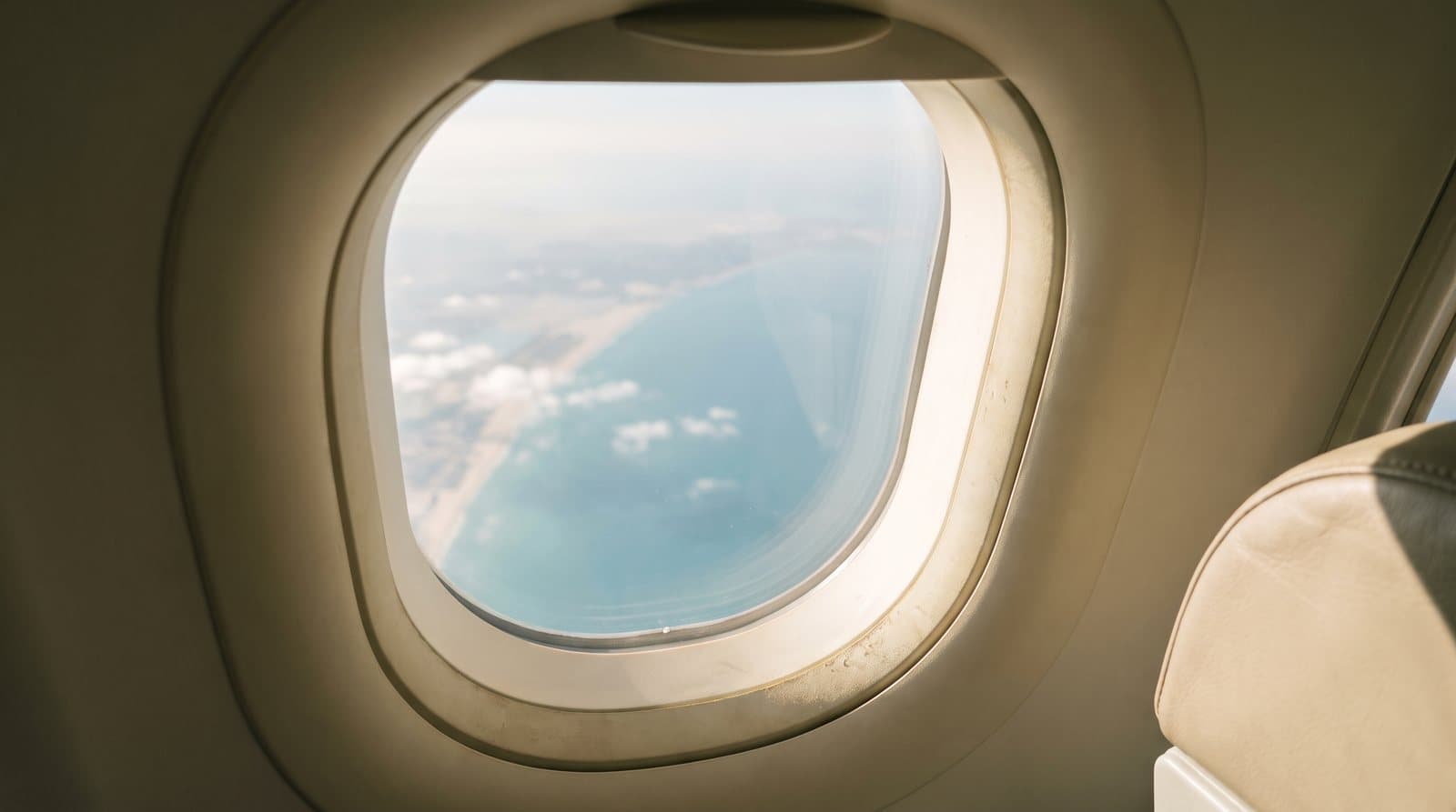 The view from an aircraft window of a turquoise sea and a pale coastline under scattered cloud.
