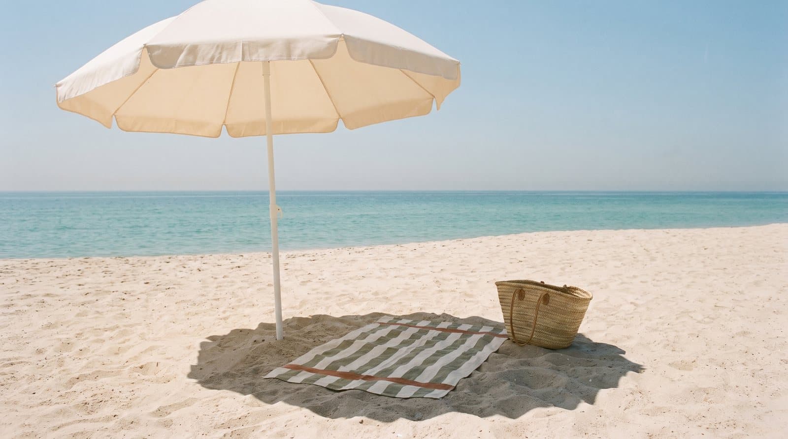 A cream beach umbrella casting a round shadow over a striped towel and a straw bag on pale sand, with a calm turquoise sea beyond.