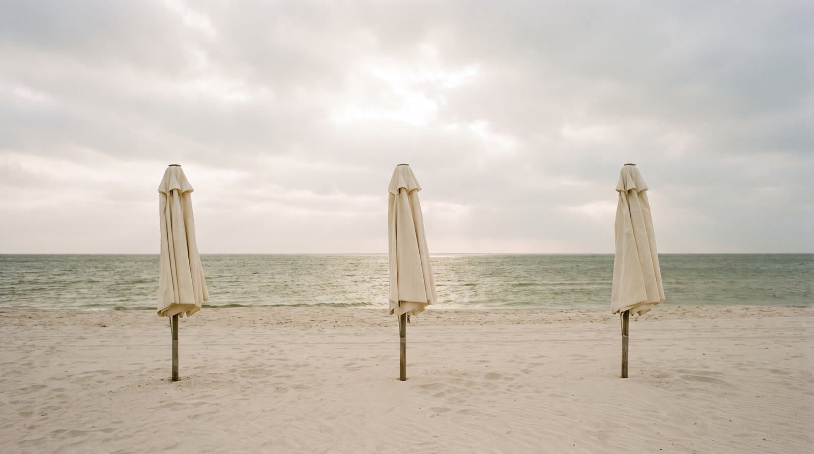Three closed cream beach umbrellas on an empty beach under a bright, overcast sky.