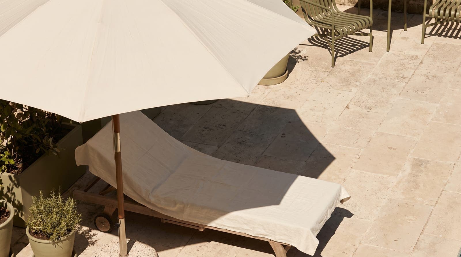 An empty linen-draped lounger beside a cream canvas umbrella on a sunlit stone terrace.