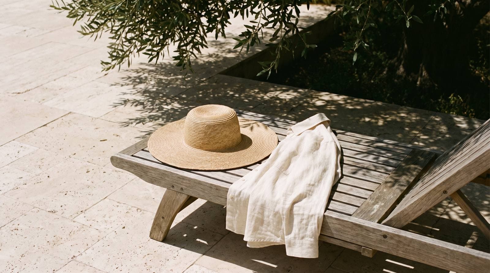 A straw sun hat and a long-sleeved linen shirt on a lounger beneath an olive tree.
