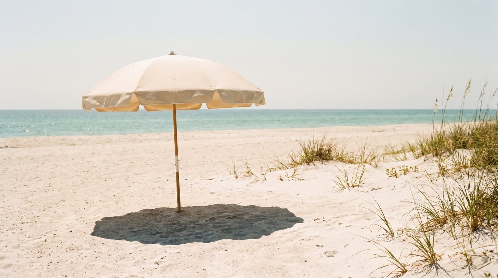 A beach umbrella's sharp midday shadow on pale sand, with the sea behind.