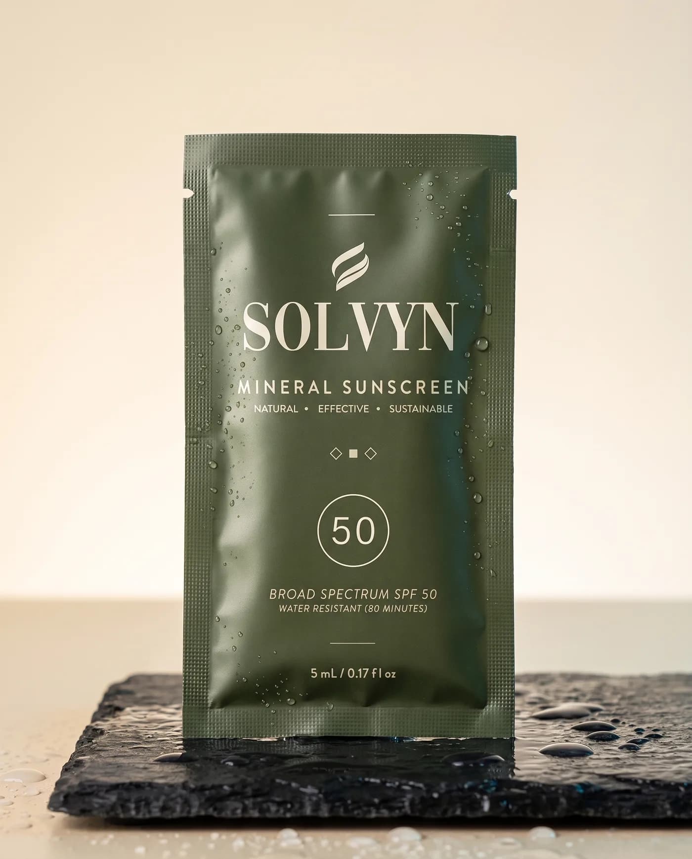 Solvyn mineral sunscreen sachet standing on wet basalt slate, beaded with water