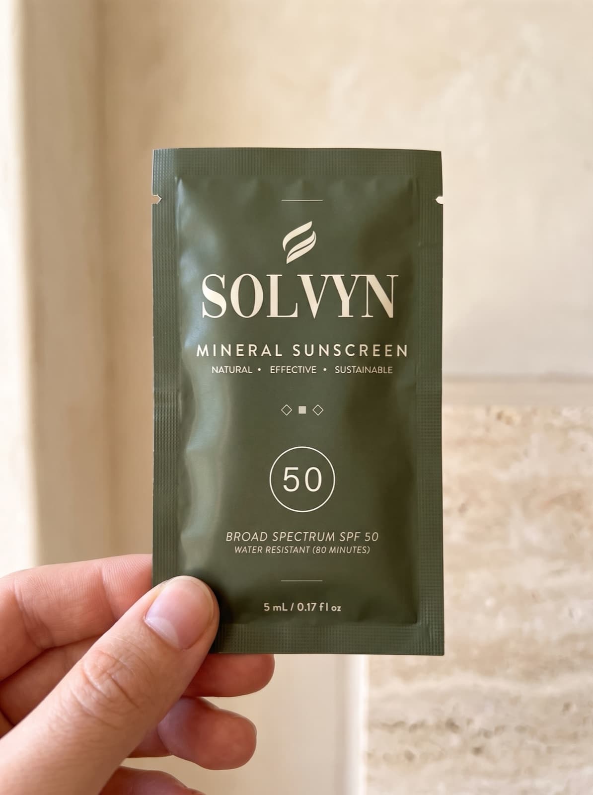 A hand holding the Solvyn Shield sachet face-on: deep olive foil printed with the Solvyn emblem, SOLVYN wordmark, MINERAL SUNSCREEN, a ring reading 50, BROAD SPECTRUM SPF 50 and WATER RESISTANT (80 MINUTES)
